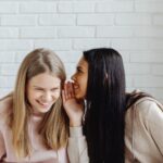 Dwie młode kobiety szepczą sobie coś do ucha i śmieją się